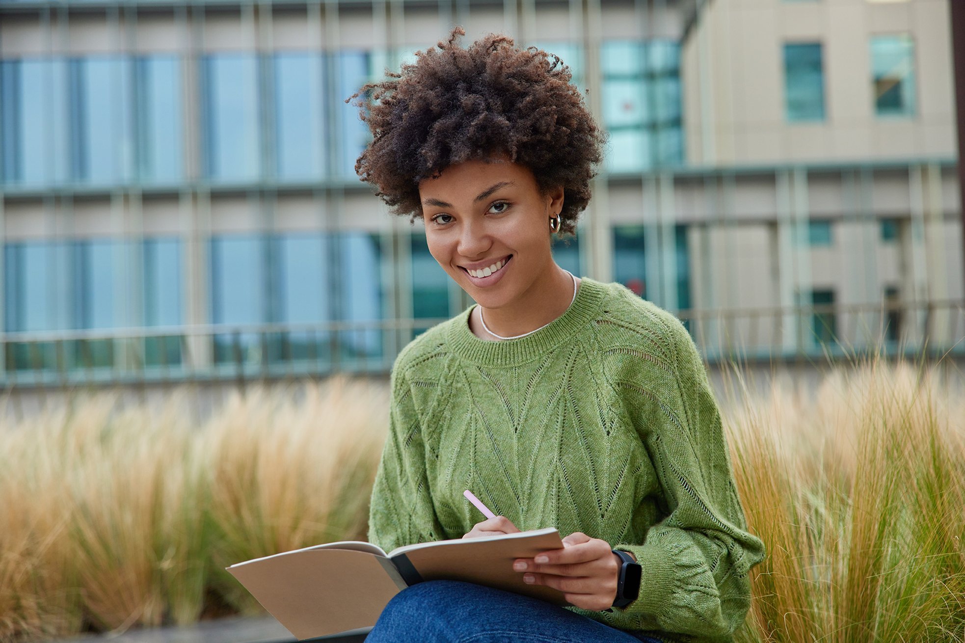 A young woman with curly hair is sitting outdoors, writing in a notebook. She is wearing a green sweater and smiling. In the background, there are modern buildings and tall grasses.
