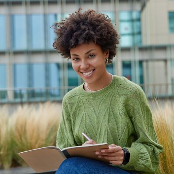 Eine junge Frau mit lockigem Haar sitzt im Freien und schreibt in ein Notizbuch. Sie trägt einen grünen Pullover und lächelt. Im Hintergrund sind moderne Gebäude und hohe Gräser zu sehen.