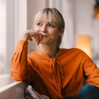Eine lächelnde Frau mit blonden Haaren sitzt am Fenster, trägt eine orangefarbene Bluse und schaut nachdenklich. Weiches Licht fällt auf ihr Gesicht, während sie mit der Hand ihr Kinn stützt.