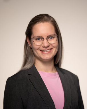 Eine lächelnde Frau mit Brille trägt einen schwarzen Blazer und ein pinkes Oberteil. Der Hintergrund ist neutral und hell.