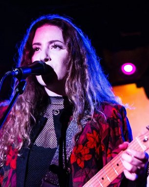A musician with curly hair stands at the microphone playing guitar. She is wearing a black top with a floral pattern. In the background, lights and another musician can be seen.