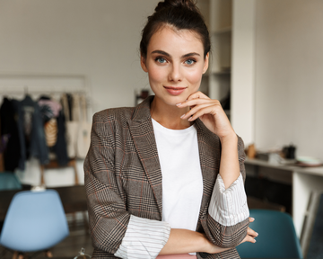 Eine junge Frau mit braunem kariertem Blazer und weißem T-Shirt steht in einem modernen Raum. Sie hat einen selbstbewussten Ausdruck und hält eine Hand an ihrem Kinn. Im Hintergrund sind Möbel und Kleidung sichtbar.