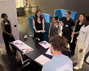 A group of people is standing around a table while participating in a presentation or discussion. A flip chart is visible in the background.