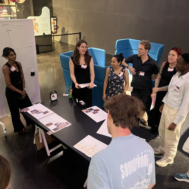 A group of people is standing around a table while participating in a presentation or discussion. A flip chart is visible in the background.
