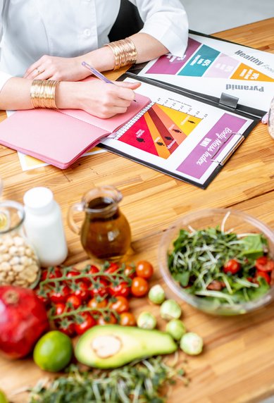 Eine Person schreibt auf einem Notizblock, umgeben von frischen Lebensmitteln wie Obst, Gemüse, Eiern und Fleisch auf einem Holztisch.