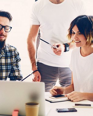 Eine Gruppe von fünf Personen sitzt um einen Tisch und schaut auf einen Laptop. Sie scheinen an einem Projekt zu arbeiten und diskutieren, während Notizblöcke und Stifte auf dem Tisch liegen.