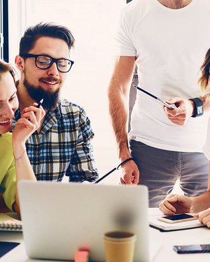 Eine Gruppe von fünf Personen sitzt um einen Tisch und schaut auf einen Laptop. Sie scheinen an einem Projekt zu arbeiten und diskutieren, während Notizblöcke und Stifte auf dem Tisch liegen.