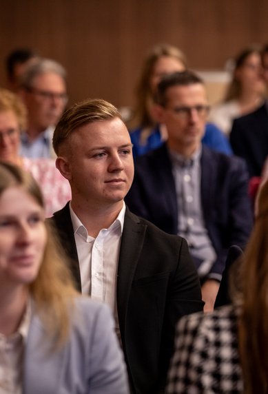 Eine Gruppe von Menschen sitzt in einem Konferenzraum, einige hören aufmerksam zu, während andere in Gespräche vertieft sind.