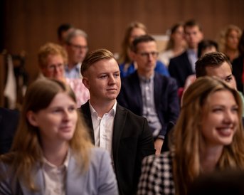 Eine Gruppe von Menschen sitzt in einem Konferenzraum, einige hören aufmerksam zu, während andere in Gespräche vertieft sind.