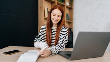 A smiling woman with long red hair is sitting at a desk, writing in a notebook with a laptop in front of her.