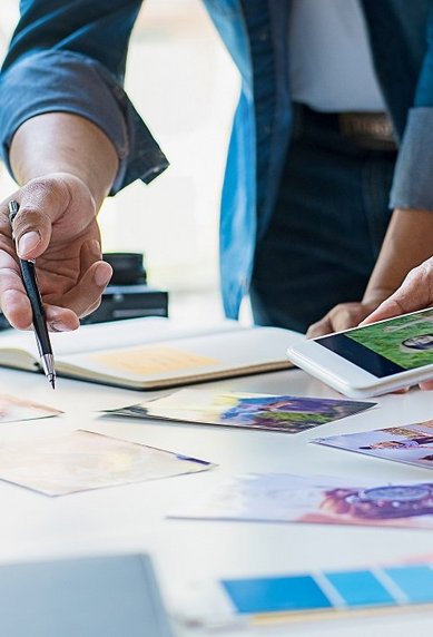 Zwei Personen arbeiten an einem Tisch, umgeben von Farbmusterkarten und Bildern. Eine Person hält ein Smartphone, während die andere mit einem Stift Notizen macht. Ein Laptop und ein Kaffeebecher sind ebenfalls sichtbar.