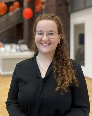 Eine lächelnde Frau mit lockigem, braunem Haar und Brille trägt ein schwarzes Oberteil. Im Hintergrund sind orangefarbene Dekorationen sichtbar.