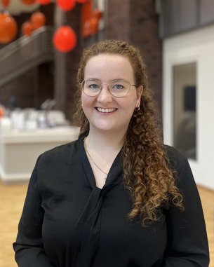 A smiling woman with curly brown hair and glasses is wearing a black top. In the background, blurred red decorations are visible.