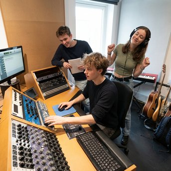 Three people in a recording studio, one person at the mixing console while the others work on a computer and enjoy the music.