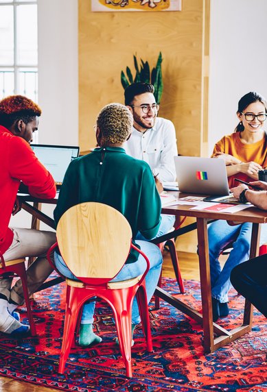 Five people are sitting at a table, working on laptops and discussing. The room is bright and furnished in a modern style.