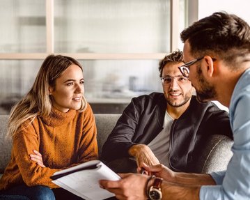 Zwei Personen sitzen auf einem Sofa und unterhalten sich mit einem dritten, der Notizen macht. Die Frau trägt einen orangefarbenen Pullover, während die anderen beiden lässige Kleidung tragen. Alle scheinen an dem Gespräch interessiert zu sein.