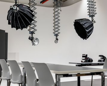 A modern photography studio with a table, white chairs, and hanging light sources. In the background, cameras and other photography equipment are visible.