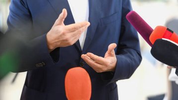 Ein Mann in einem Anzug spricht mit gestikulierenden Händen vor Mikrofonen in einem Presseinterview.