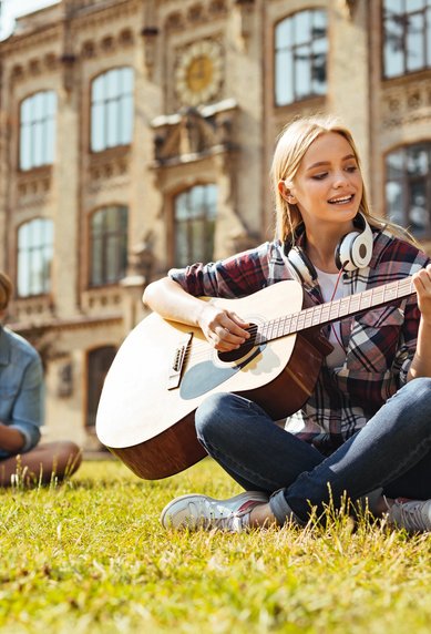 A young woman is playing guitar on a meadow while two men in the background are working on a laptop and a book.