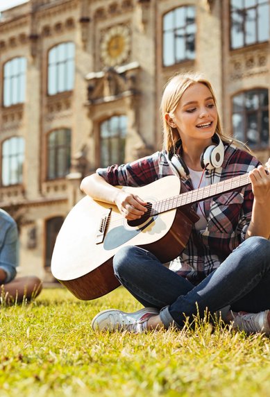 Eine junge Frau spielt Gitarre auf einer Wiese, während zwei Männer im Hintergrund an einem Laptop und einem Buch arbeiten.