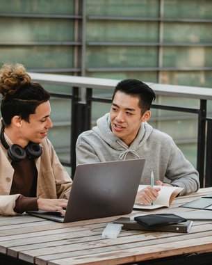 Zwei junge Männer sitzen an einem Tisch im Freien und arbeiten zusammen an einem Laptop. Der Mann links trägt Kopfhörer und hat einen entspannten Ausdruck, während der Mann rechts Notizen macht und auf den Bildschirm schaut.