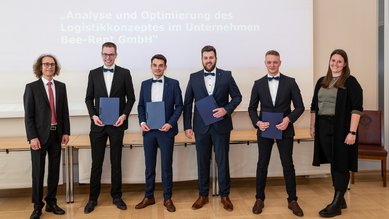 Sechs Personen in formeller Kleidung stehen zusammen, halten Urkunden in den Händen. Im Hintergrund ist ein Präsentationsbildschirm sichtbar.