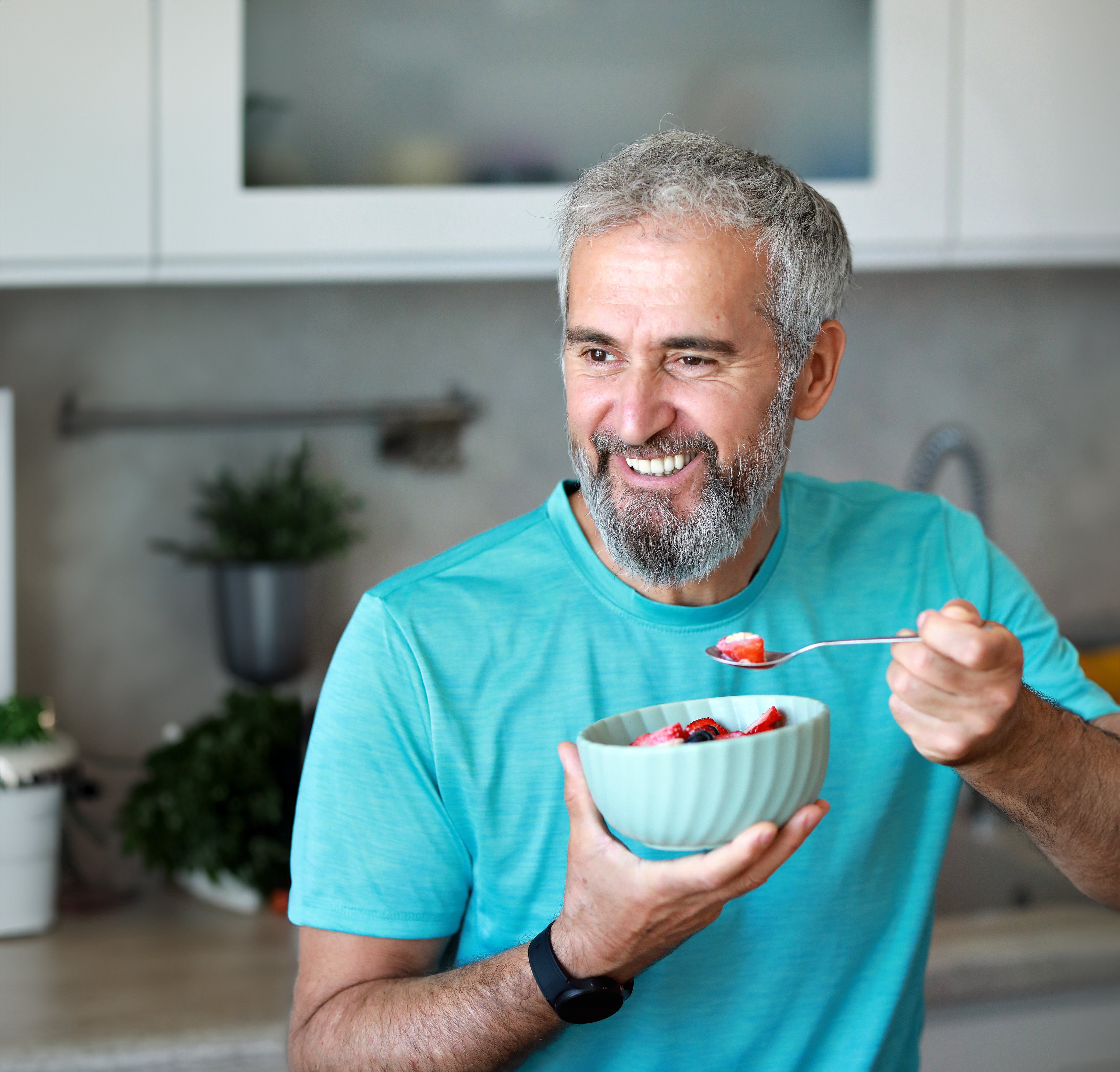 A smiling man with a gray beard holds a bowl of fruit and scoops some out while standing in a modern kitchen.