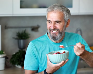 Ein lächelnder Mann mit grauem Bart hält eine Schüssel mit Obst und löffelt davon, während er in einer modernen Küche steht.