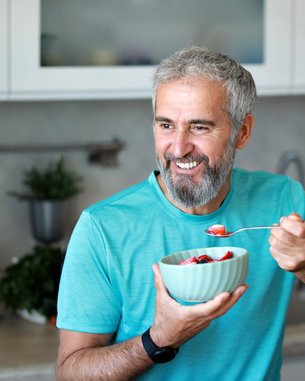Ein lächelnder Mann mit grauem Bart hält eine Schüssel mit Obst und löffelt davon, während er in einer modernen Küche steht.