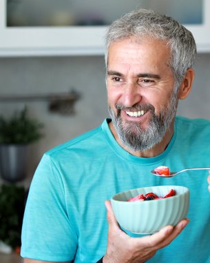 A smiling man with a gray beard holds a bowl of fruit and scoops some out while standing in a modern kitchen.