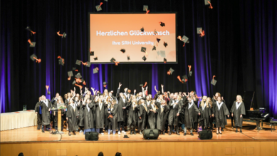 Graduates of SRH University celebrate their graduation as they toss their caps into the air. A banner congratulates them.
