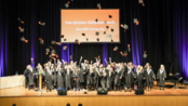 Graduates of SRH University celebrate their graduation as they toss their caps into the air. A banner congratulates them.