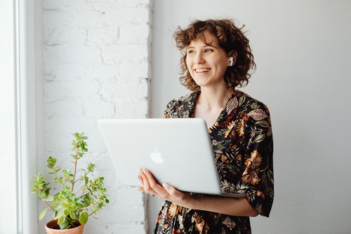 A smiling woman with curly hair is holding a laptop in her hand. She is wearing a floral top and standing next to a potted plant. The background features a bright, brick wall.
