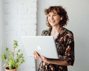 A smiling woman with curly hair is holding a laptop in her hand. She is wearing a floral top and standing next to a potted plant. The background features a bright, brick wall.