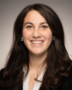 Eine lächelnde Frau mit langen, dunklen Haaren trägt eine schwarze Jacke und eine helle Bluse. Der Hintergrund ist neutral und unscharf.