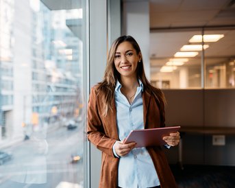 A smiling woman stands by a window in a modern office building, holding a tablet in her hand. In the background, urban buildings and a street are visible.