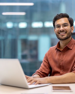 Ein Mann mit Brille sitzt an einem Schreibtisch und arbeitet an einem Laptop. Er trägt ein rotes Hemd und lächelt. Auf dem Tisch befinden sich ein Stiftbehälter und ein Notizbuch.