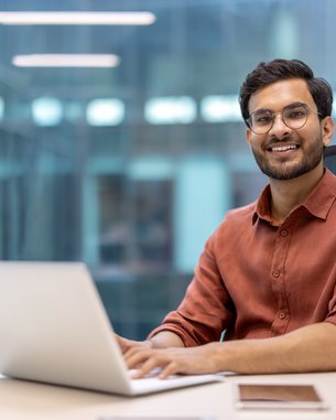 Ein Mann mit Brille sitzt an einem Schreibtisch und arbeitet an einem Laptop. Er trägt ein rotes Hemd und lächelt. Auf dem Tisch befinden sich ein Stiftbehälter und ein Notizbuch.
