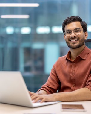 Ein Mann mit Brille sitzt an einem Schreibtisch und arbeitet an einem Laptop. Er trägt ein rotes Hemd und lächelt. Auf dem Tisch befinden sich ein Stiftbehälter und ein Notizbuch.