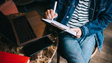 A person is sitting at a table, writing on a notepad, with a laptop and a smartphone in front of them.
