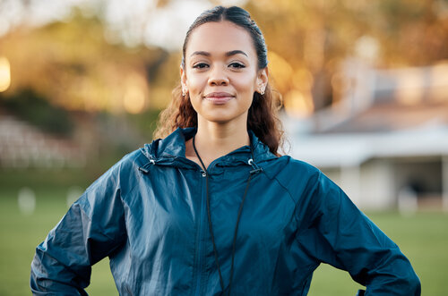 A young woman with curly hair is wearing a sports jacket and stands confidently outdoors, with a neutral expression.