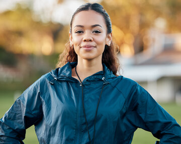 A young woman with curly hair is wearing a sports jacket and stands confidently outdoors, with a neutral expression.