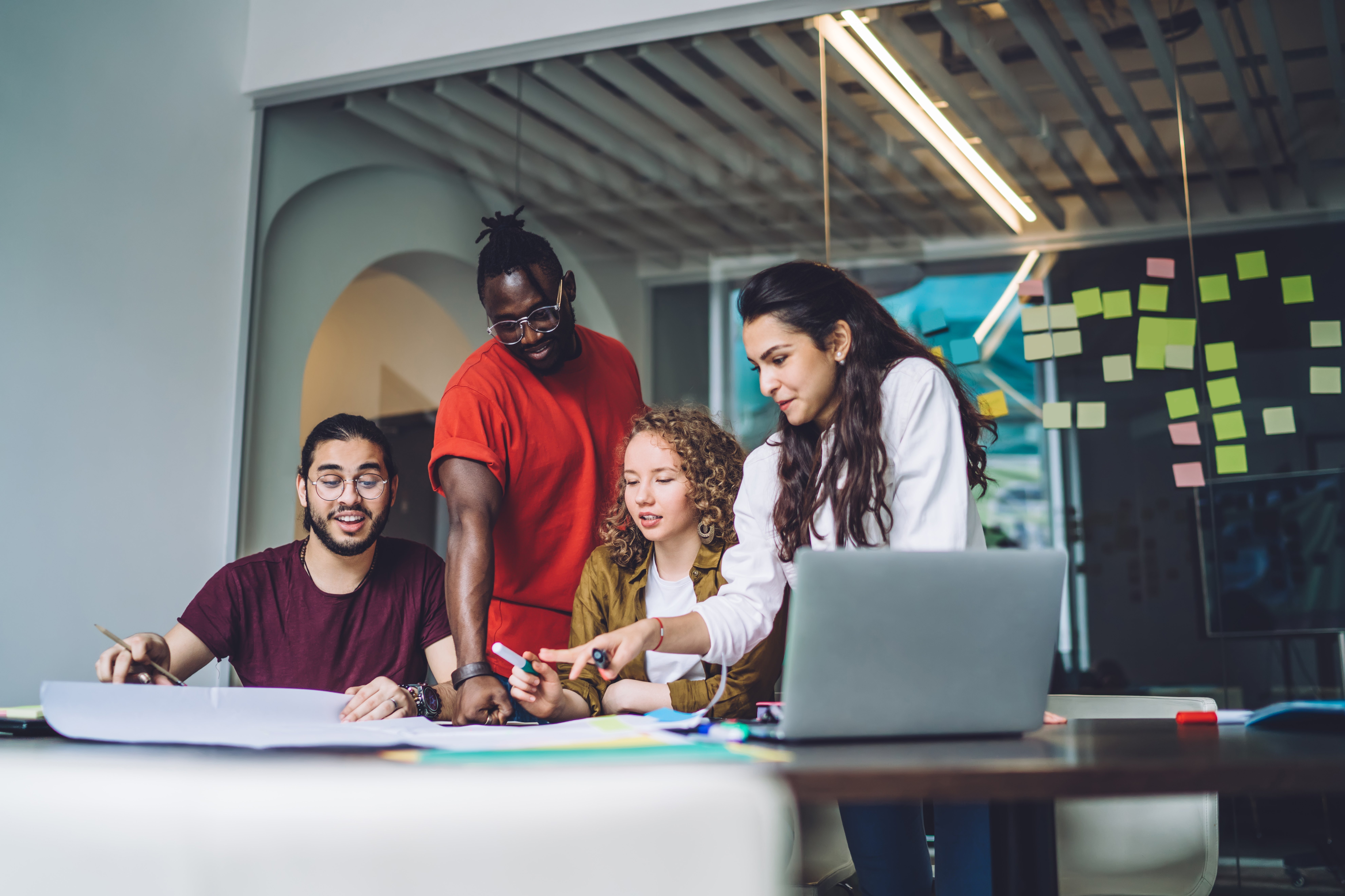 Four people are working together at a table, surrounded by notes and a laptop. They are discussing and sketching ideas on a large sheet of paper. The atmosphere feels collaborative and creative.