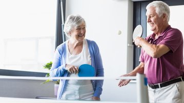 An older couple is playing table tennis in a bright room. The woman is holding a paddle and smiling, while the man hits the ball.