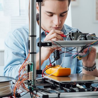 A young man is focused on a 3D printer, checking cables and components. In the background, another man can be seen with a laptop.