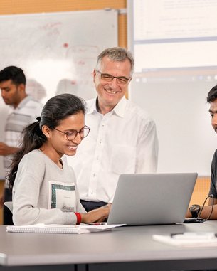 A group of students is working on laptops in a modern classroom. A professor smiles and watches a student who is working on her computer, while other students stand in the background.