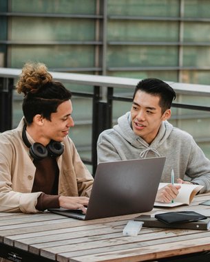 Zwei junge Männer sitzen an einem Tisch im Freien. Einer arbeitet an einem Laptop, während der andere Notizen macht. Beide scheinen an einem Projekt oder einer Diskussion beteiligt zu sein.