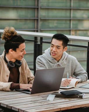 Zwei junge Männer sitzen an einem Tisch im Freien. Einer arbeitet an einem Laptop, während der andere Notizen macht. Beide scheinen an einem Projekt oder einer Diskussion beteiligt zu sein.