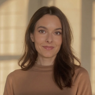 A woman with long brown hair is wearing a brown top and is smiling slightly in a bright room.