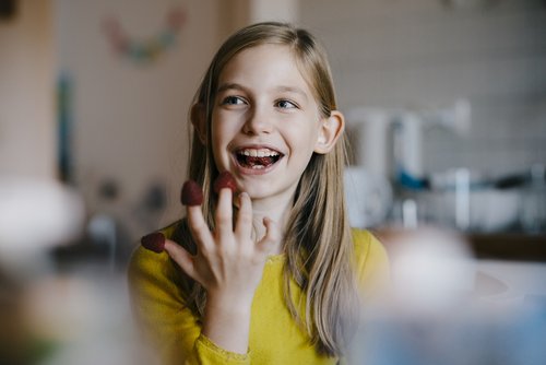 Ein Mädchen mit langen Haaren trägt ein gelbes Oberteil und lächelt, während es Erdbeeren auf ihren Fingern hält. Im Hintergrund sind verschwommene Küchenutensilien zu sehen.